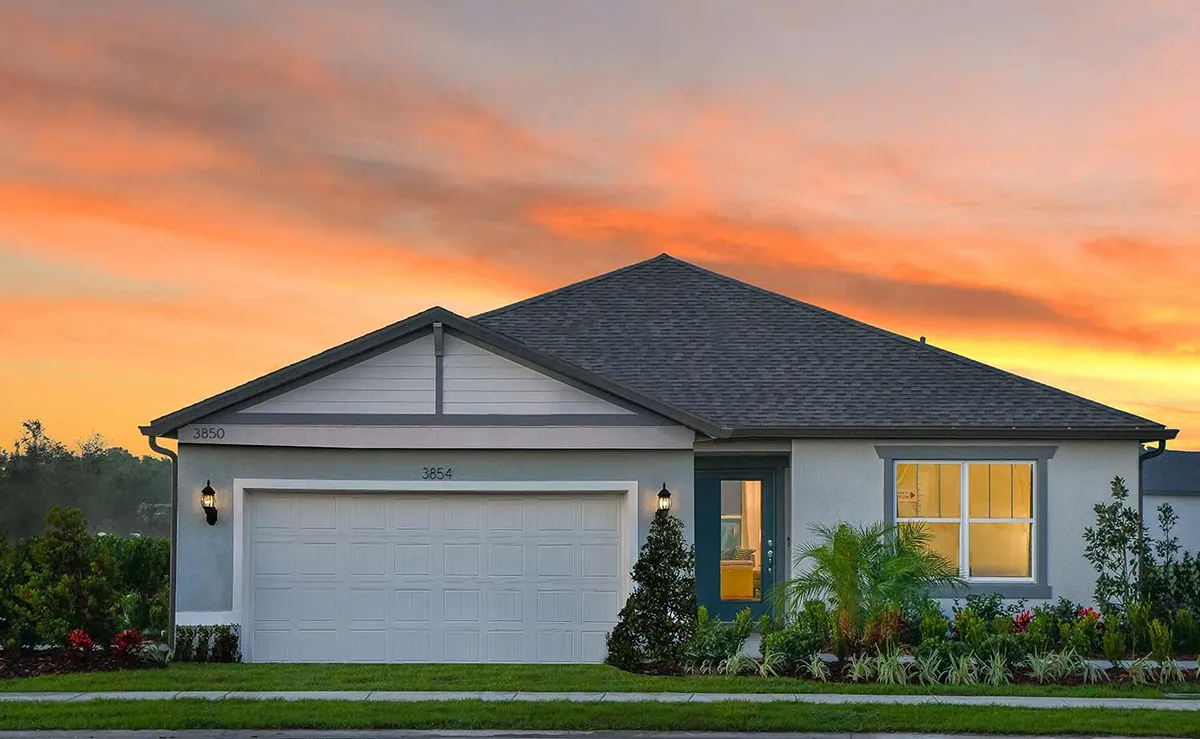 Single-story house with gray roof, white garage door, and blue front door at sunset. Warm light glows from a window, with green shrubs and landscaping in front. The sky is streaked with orange and yellow hues.