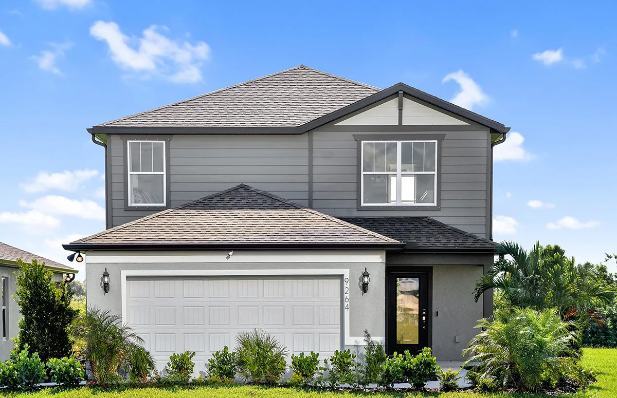 Two-story modern suburban house with gray siding, white trim, and a double garage. There are large front windows, a central front door, and well-maintained landscaping with green bushes and palm plants. The sky is clear and blue.