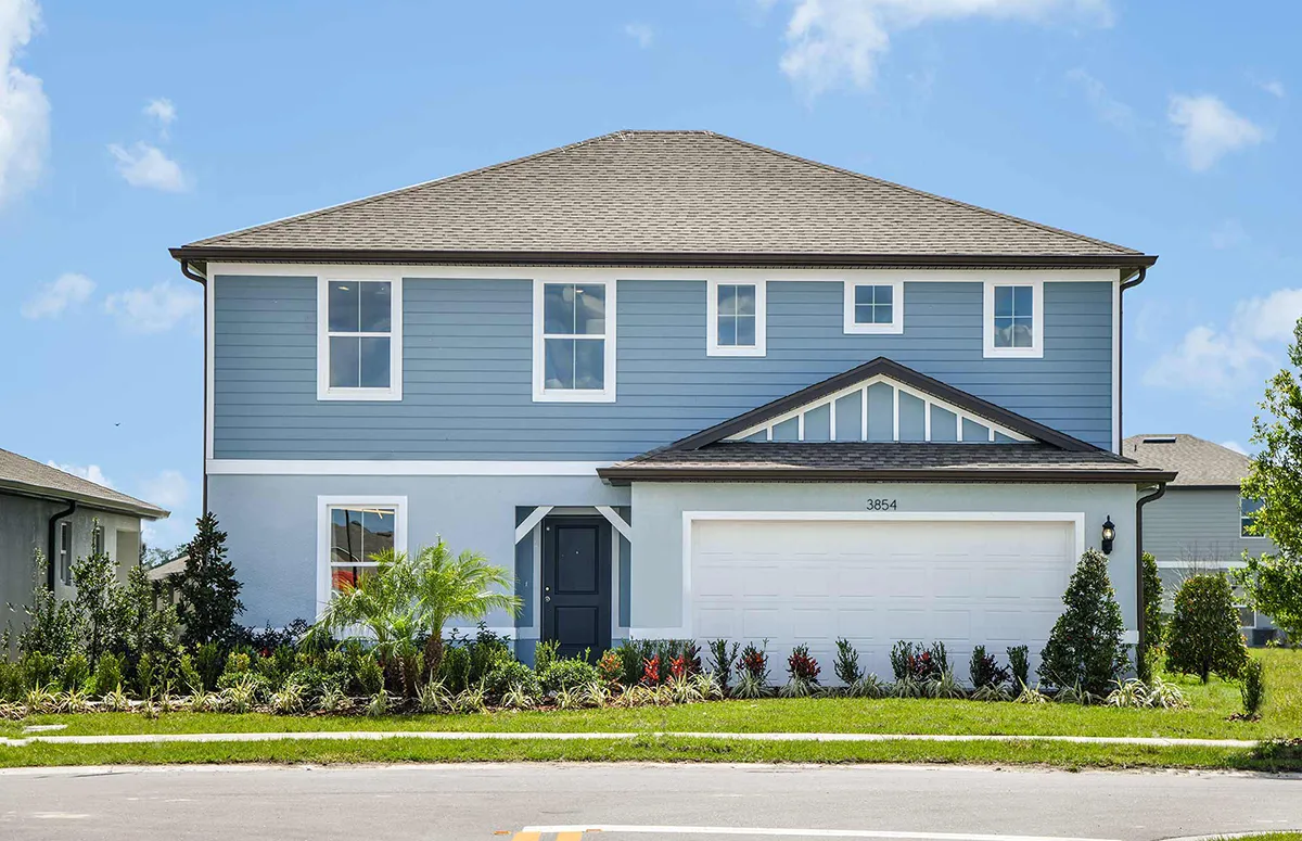 A two-story blue house with white trim, a gray shingled roof, and a white double garage door. There are shrubs and palm plants in the landscaped front yard, and the sky is clear with a few clouds.