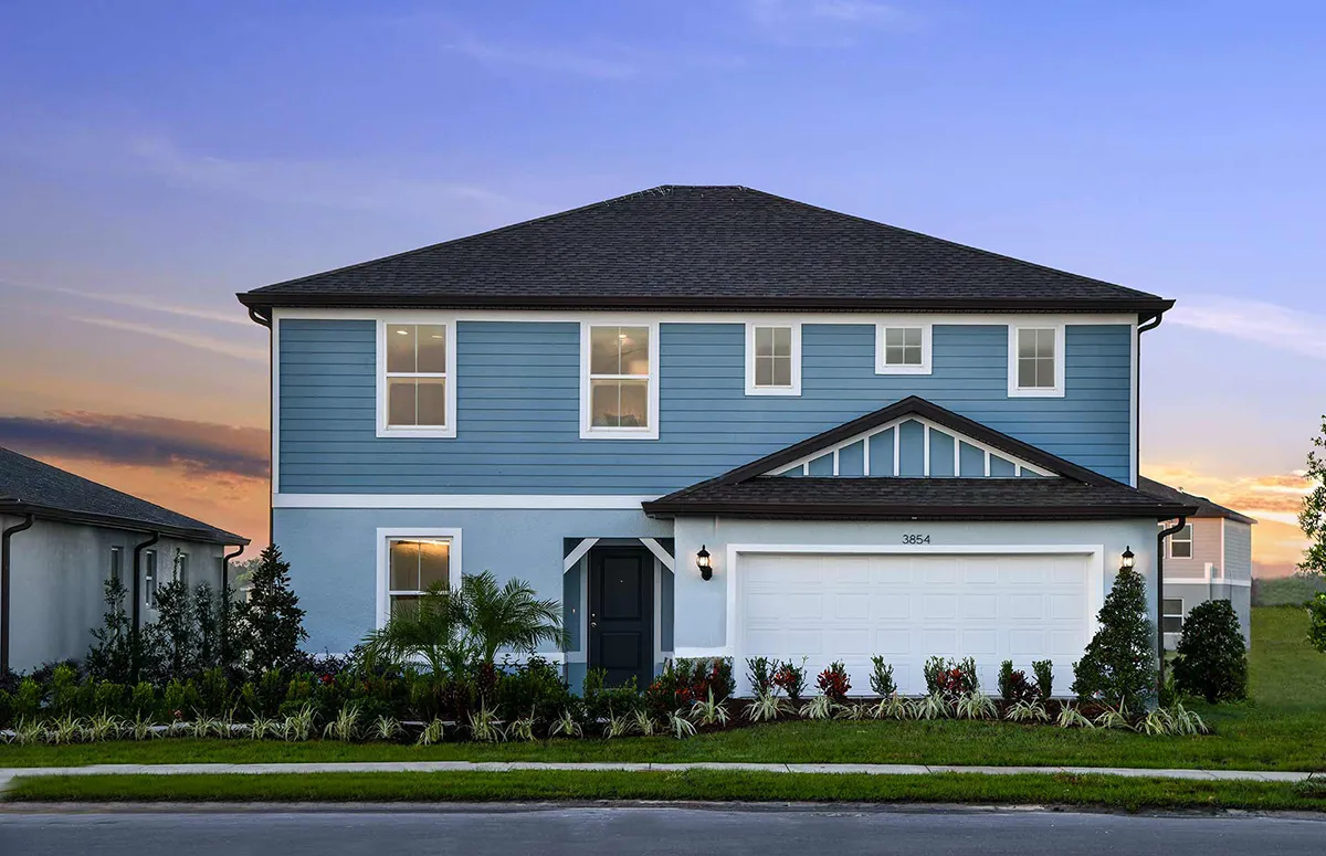 A two-story blue house with white trim and a dark roof, featuring a front porch, double garage, and landscaped yard, photographed at dusk with a colorful sunset sky in the background.