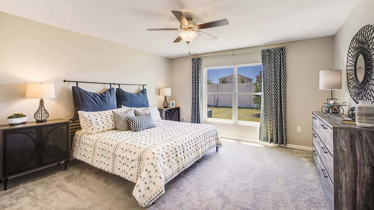 A bright bedroom with a large bed, patterned bedding, bedside tables with lamps, a ceiling fan, a dresser with decor, and a window with curtains letting in natural light.