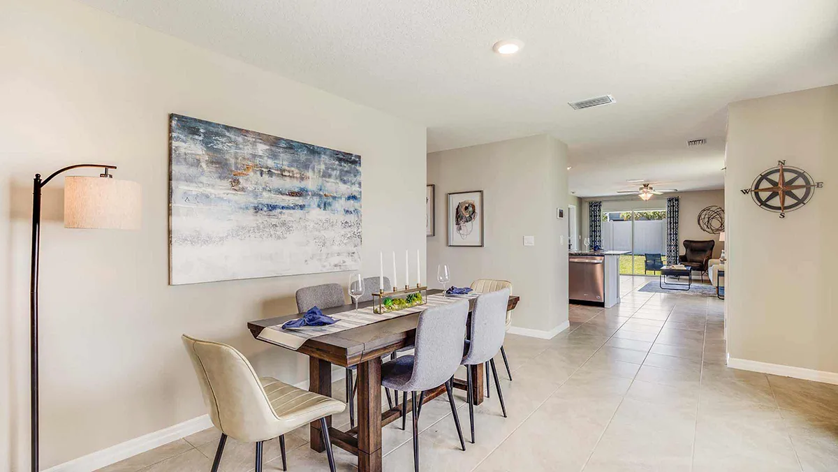 A modern dining area with a wooden table set for four, surrounded by mixed gray and beige chairs. Neutral walls, abstract wall art, and a standing lamp enhance the bright, open space leading to a living room and kitchen.
