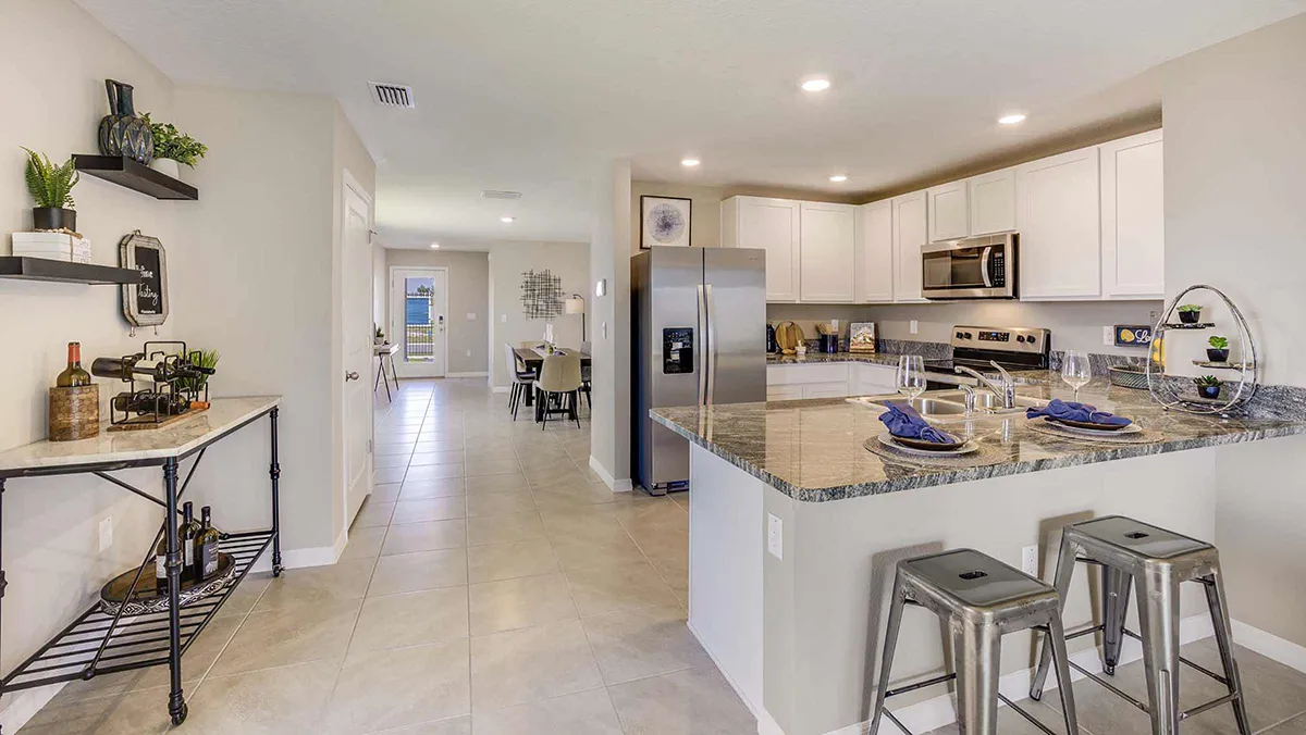 Modern kitchen with white cabinets, stainless steel appliances, granite countertops, and a breakfast bar with two metal stools. A dining area is visible in the background, and decorative shelves line the left wall.