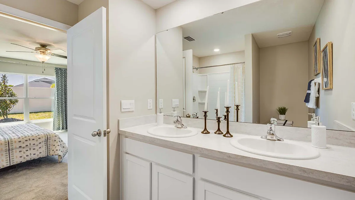 Modern bathroom with double sinks, large mirror, and decorative candlesticks on the counter; a bedroom and a window with a view of a yard are visible through an open door.