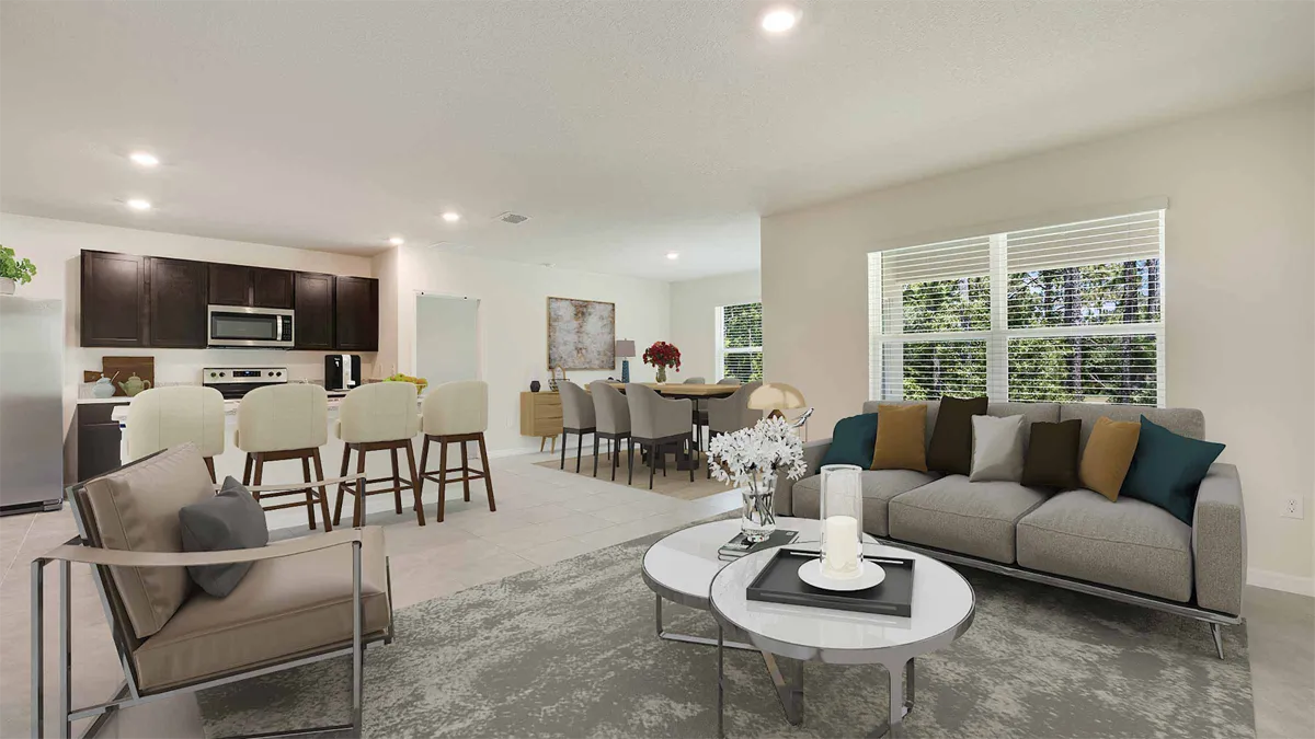 Modern open-concept living space with a gray sofa and armchair, a round coffee table, dining area with a table and chairs, and a kitchen with dark cabinets, white countertops, and a breakfast bar with stools. Large window lets in sunlight.