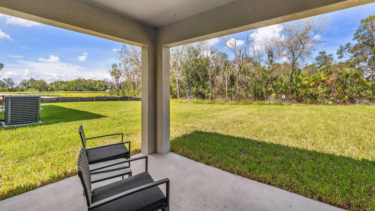 Covered patio with two black chairs overlooks a spacious green backyard with trees and blue sky in the background. An air conditioning unit is visible on the left side of the yard.