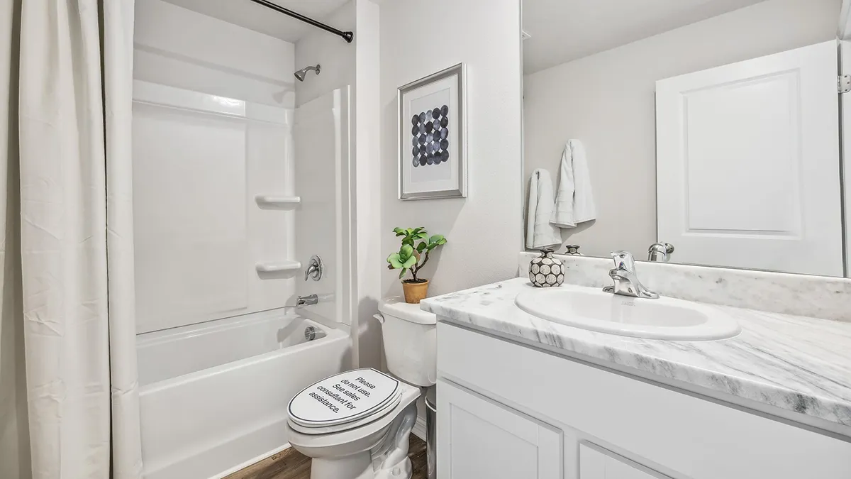 White bathroom with a bathtub and shower, white curtain, toilet with a sign on the lid, marble countertop sink, small plant, towels, and framed wall art above the toilet.