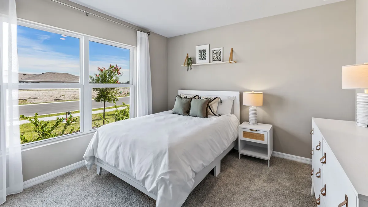 A bright, modern bedroom with a white bed, gray and green pillows, a nightstand with a lamp, a white dresser, and large window with outdoor view of a road, greenery, and hills. Neutral walls and carpeted floor.