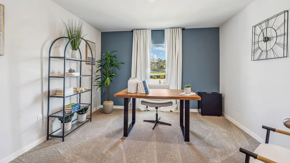 A modern home office with a wooden desk, white chair, metal bookshelf with plants and books, and large window with white curtains letting in natural light. The walls are white with one blue accent wall.