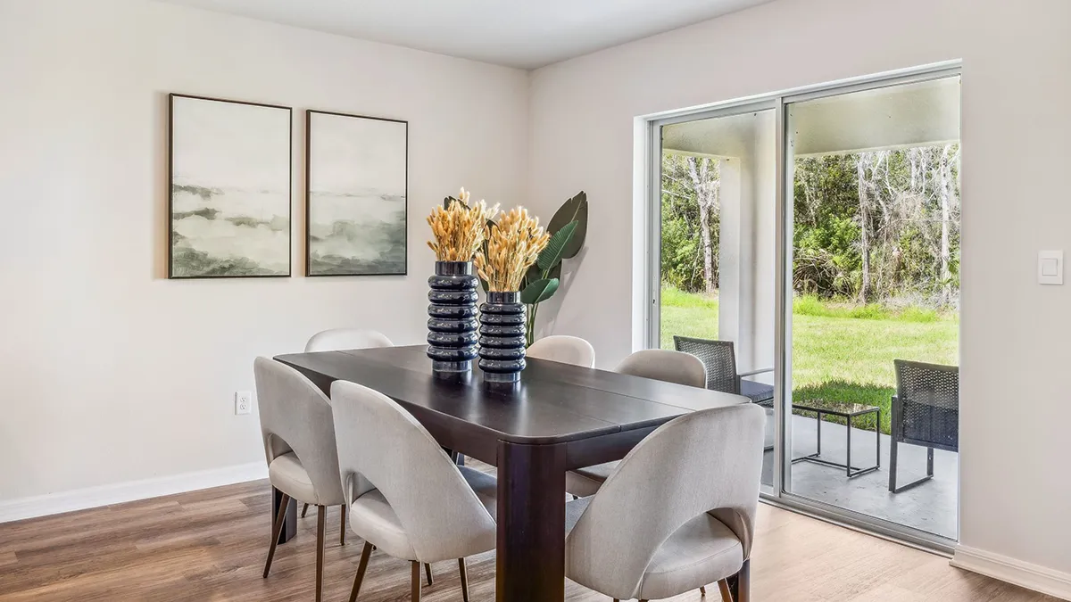 Modern dining room with a dark wooden table, six light beige chairs, two black vases with dried flowers, two abstract paintings on the wall, and sliding glass doors opening to a green outdoor area.