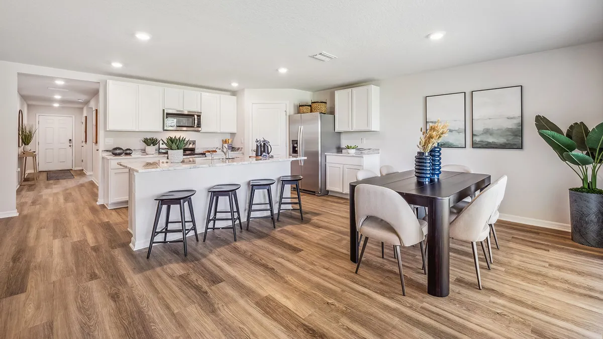 Modern kitchen and dining area with wood flooring, white cabinets, stainless steel appliances, an island with four black stools, a dark dining table with six chairs, and decorative plants and artwork.