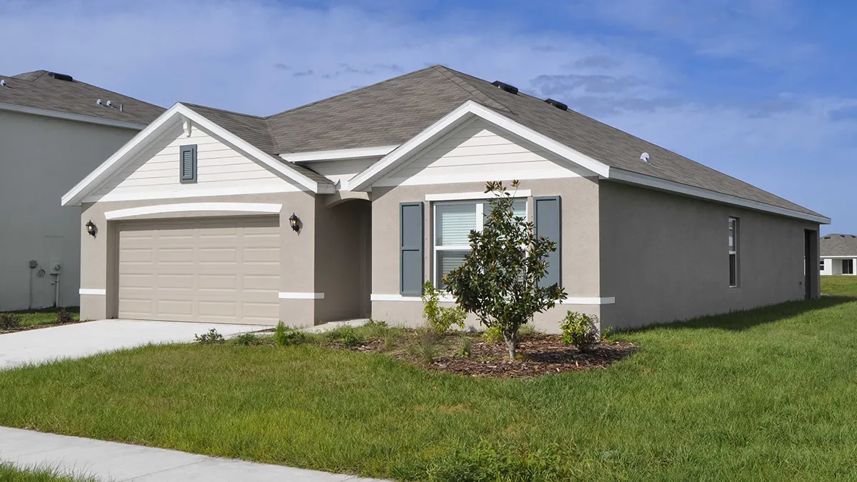 A single-story beige house with a two-car garage, white trim, and gray shutters, surrounded by green grass and a small landscaped area in the front yard under a partly cloudy sky.