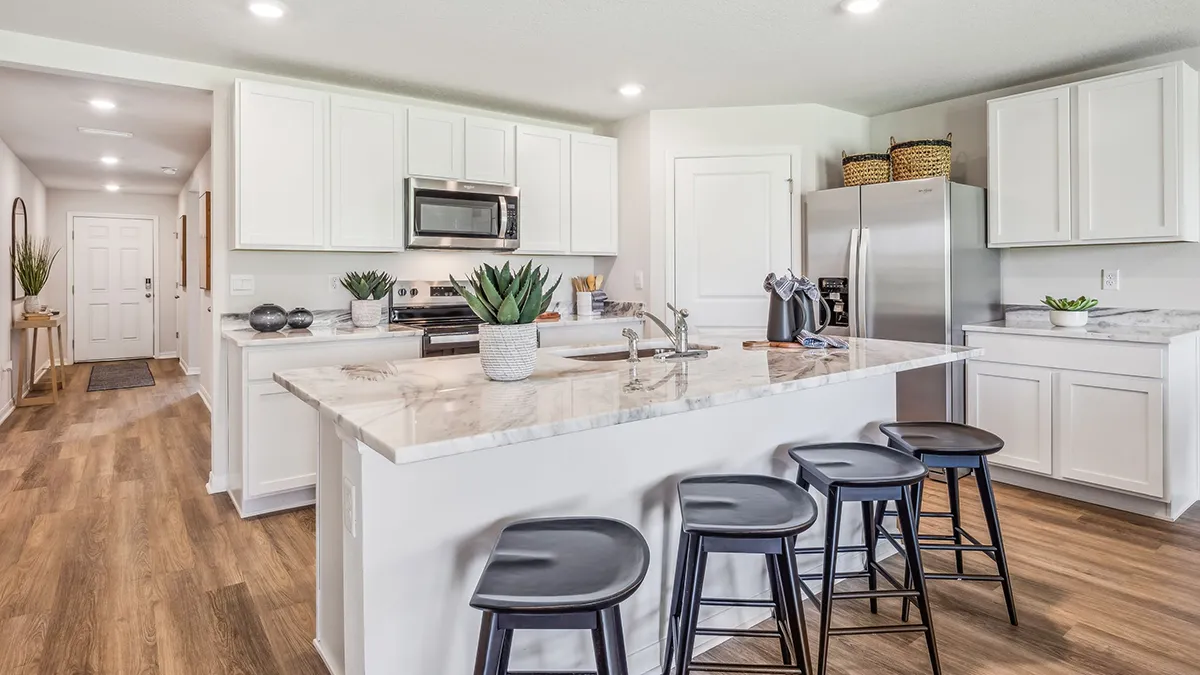 Modern kitchen with white cabinets, stainless steel appliances, a marble island with a sink, black stools, and potted plants. Wood flooring extends into a hallway with closed doors. The space is bright and tidy.