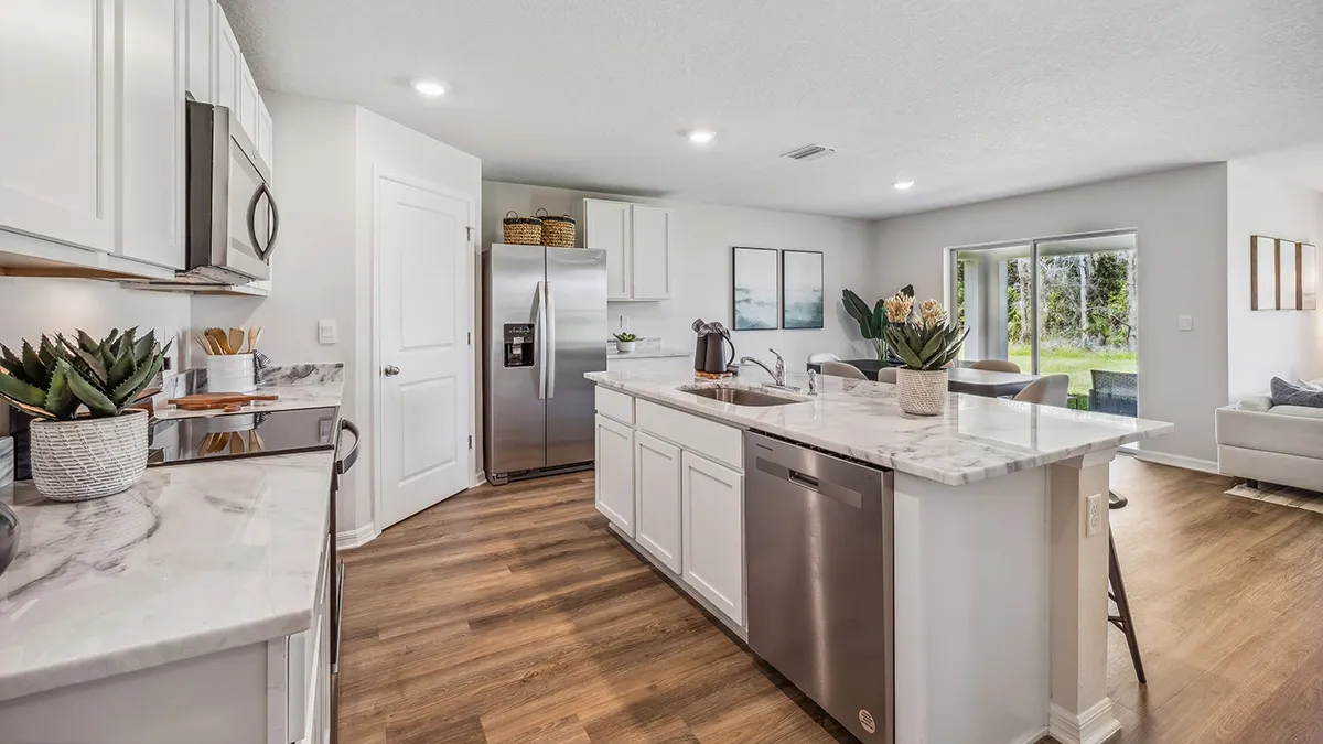Modern kitchen with white cabinets, marble countertops, stainless steel appliances, and a center island. The kitchen opens into a bright living and dining area with large windows and wood flooring throughout.