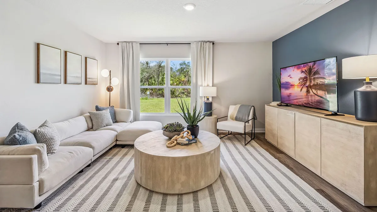 Modern living room with a white sectional sofa, round wood coffee table, striped rug, TV on a light wood console, floor lamp, large window with white curtains, and plant decor.