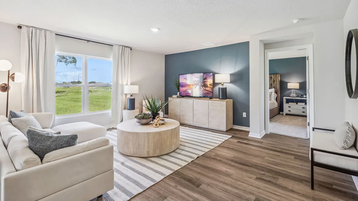 Modern living room with a white sofa, round wooden coffee table, striped rug, TV on a light wood console, and large window with outdoor view. Bedroom with neutral decor is visible through an open doorway.