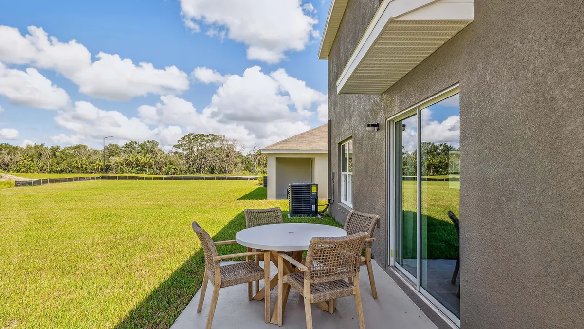 A round table with four wicker chairs sits on a concrete patio outside a house, overlooking a large green lawn under a blue sky with scattered clouds.