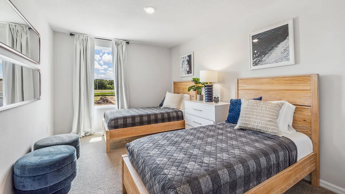 Bright bedroom with two wooden twin beds, gray bedding, a white dresser with a lamp and plant, two blue ottomans, three mirrors, and a window with white curtains letting in natural light.
