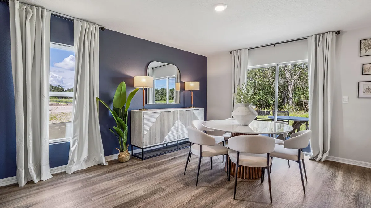 Modern dining room with wood floor, round glass table, six white chairs, large windows with white curtains, blue accent wall, sideboard with mirror and two lamps, and a tall potted plant. Trees visible outside.