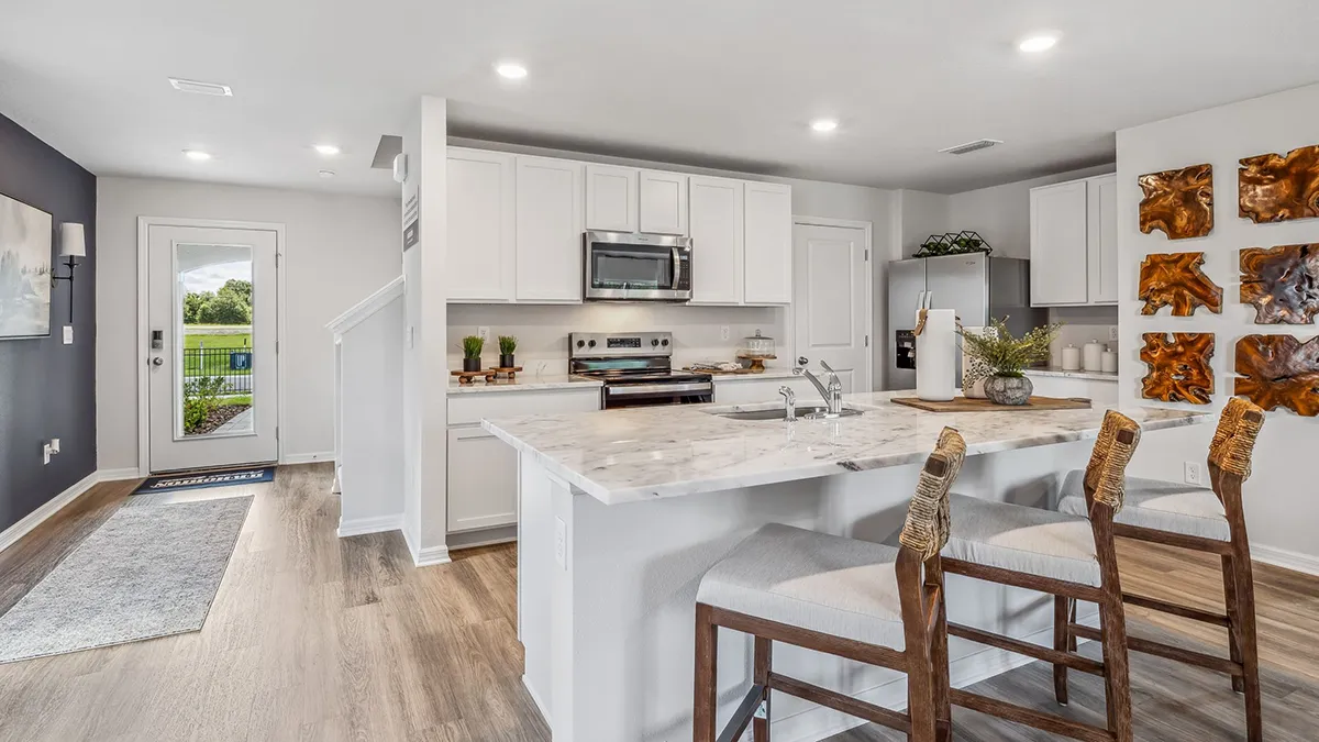Modern kitchen with white cabinets, stainless steel appliances, marble island with sink, and two bar stools. Wooden art pieces decorate the wall, and a door leads to a backyard with green grass visible outside.