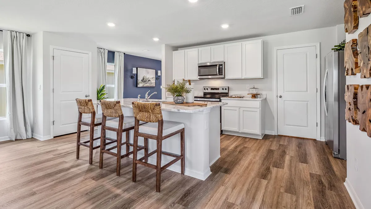 Modern kitchen with white cabinets, stainless steel appliances, and light wood flooring. An island with a white countertop features three woven barstools. Large windows and decorative wall accents provide natural light and style.