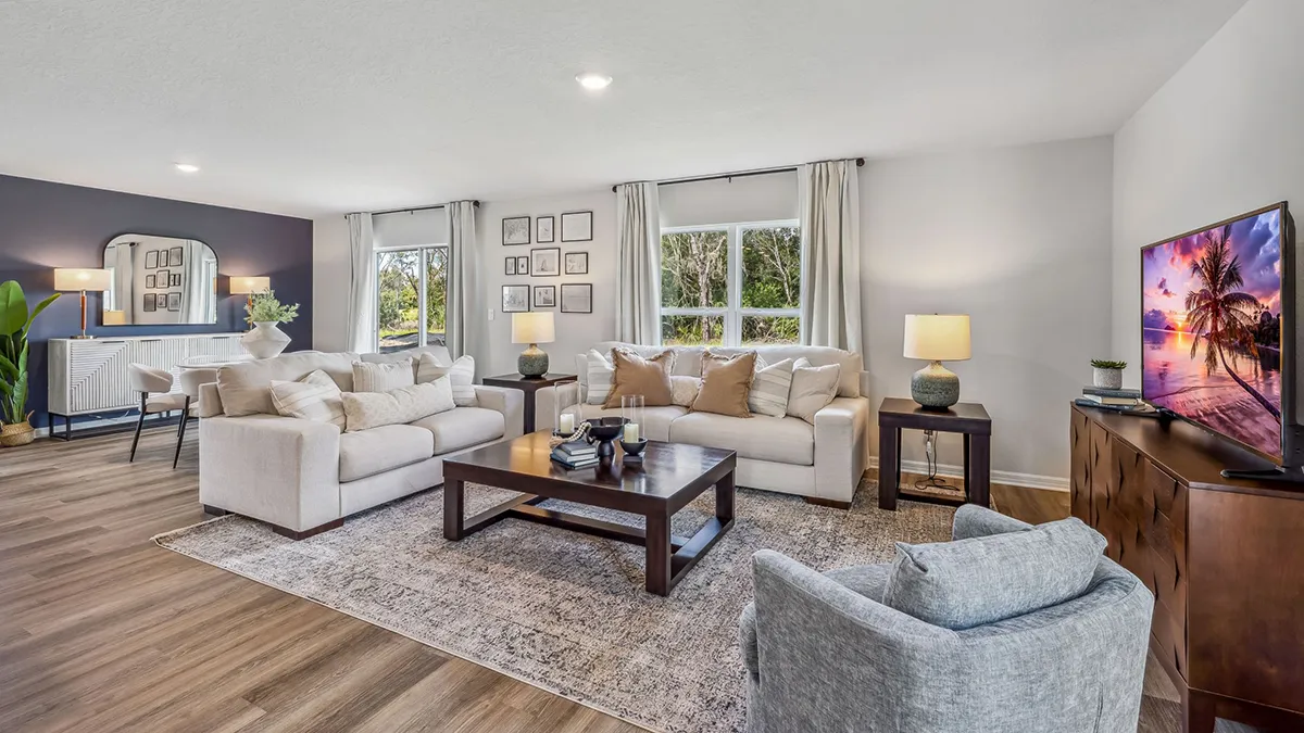 Bright, modern living room with neutral-toned sofas, a gray armchair, wooden coffee table, TV displaying a beach scene, large rug, wall art, and windows with light curtains letting in natural light.