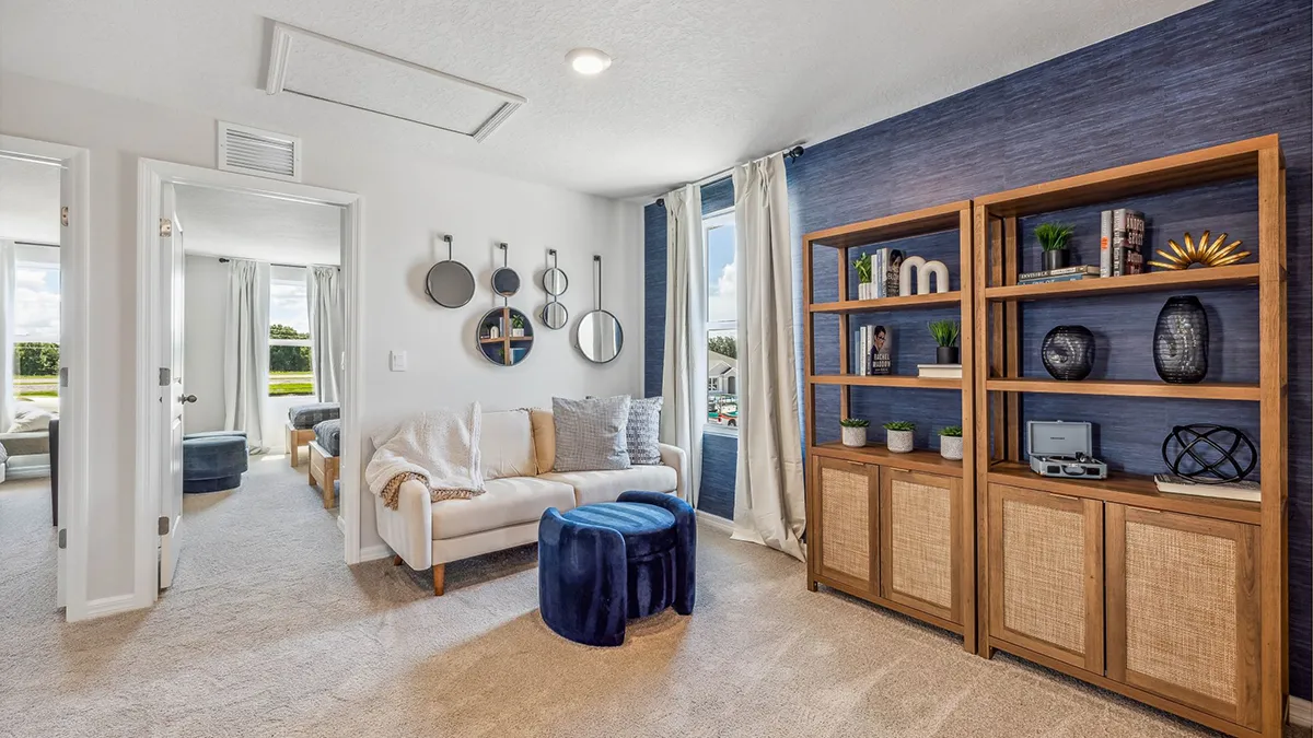 A cozy living room with a white sofa, blue ottomans, wall mirrors, large window with curtains, and wooden bookshelves with decorative items on a blue accent wall. Neutral carpet and bright natural light.