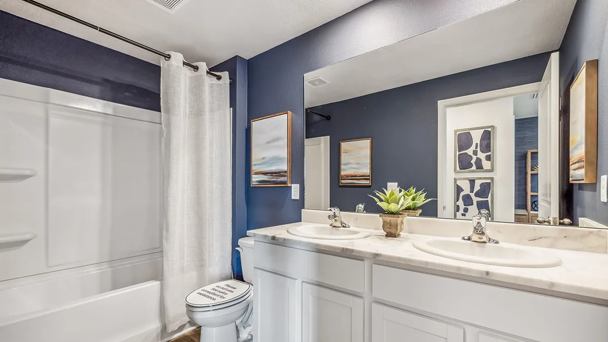 A modern bathroom with dark blue walls, a white bathtub with a shower curtain, a double sink vanity with white cabinets, a potted plant on the counter, and abstract art on the walls.