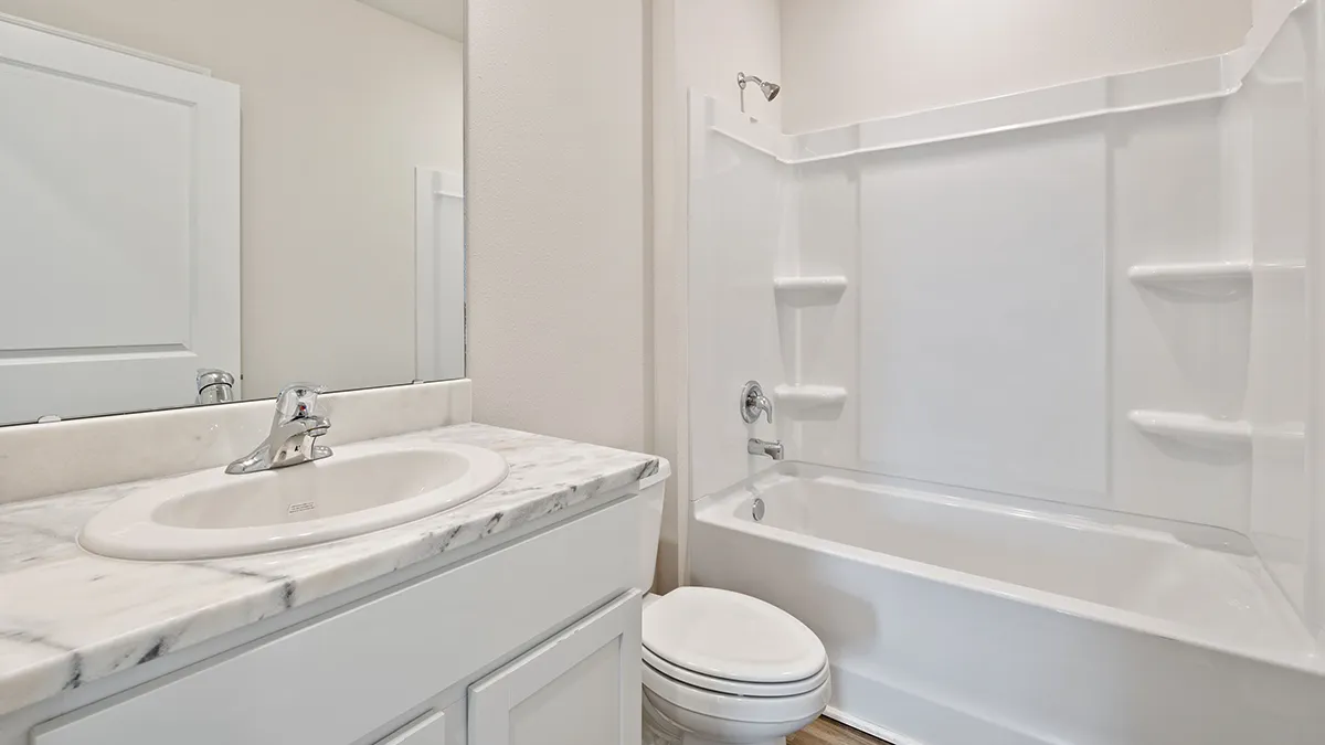 A clean, modern bathroom with a white sink and marble countertop, a white toilet, and a white bathtub with built-in shelves and a showerhead, set against light-colored walls.