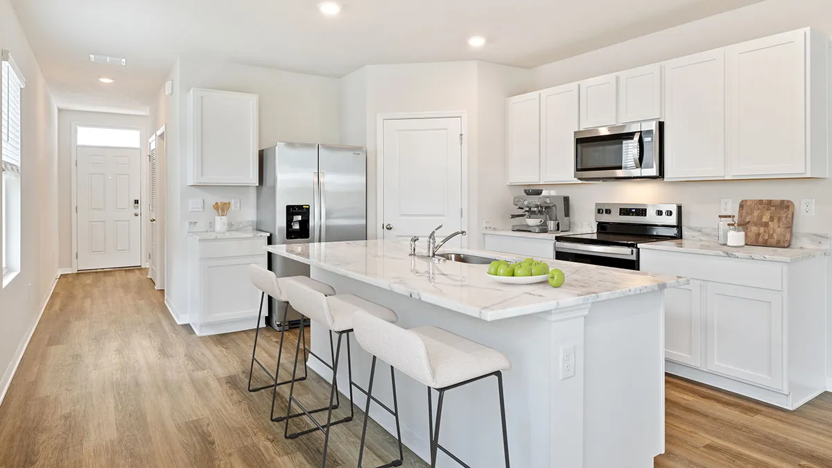 Modern kitchen with white cabinets, stainless steel appliances, a marble island countertop with three beige barstools, and light wood flooring. A bowl of green apples sits on the island. Bright and clean atmosphere.