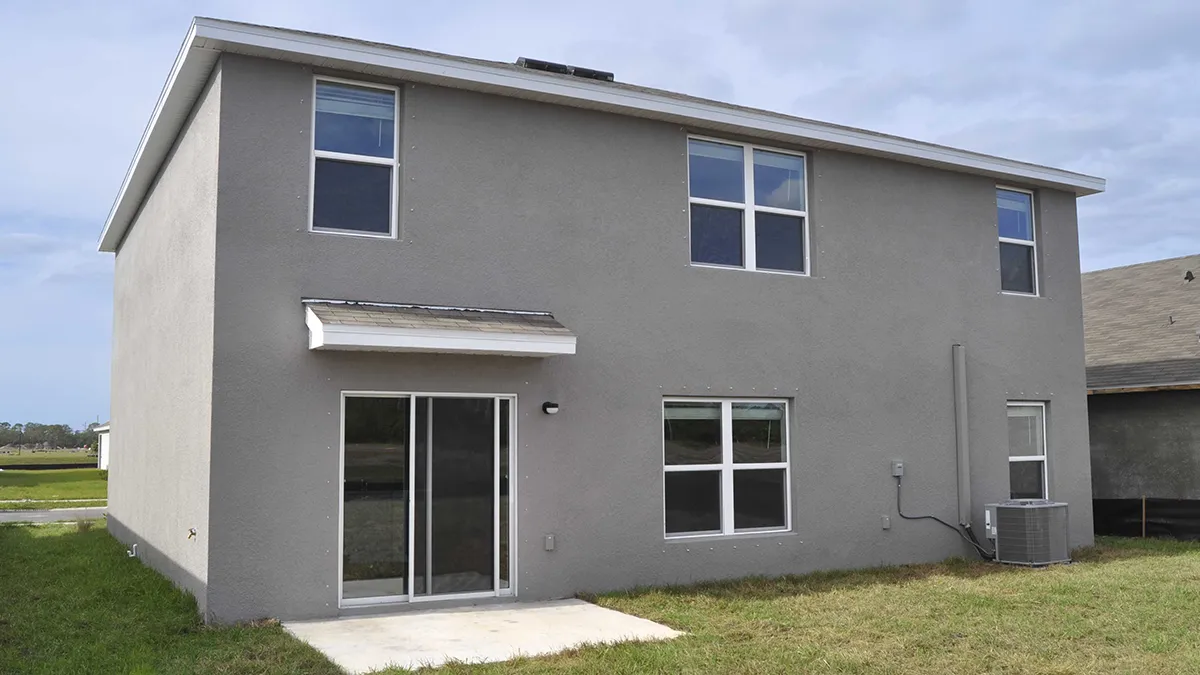A two-story gray house with four windows on the upper floor and three windows plus a sliding glass door on the lower floor, opening to a small concrete patio in a grassy backyard.