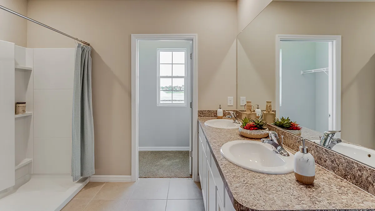 Modern bathroom with a double sink vanity, granite countertop, and large mirror. To the left is a walk-in shower with a curtain. A doorway leads to a room with a window and closet. Neutral color palette throughout.
