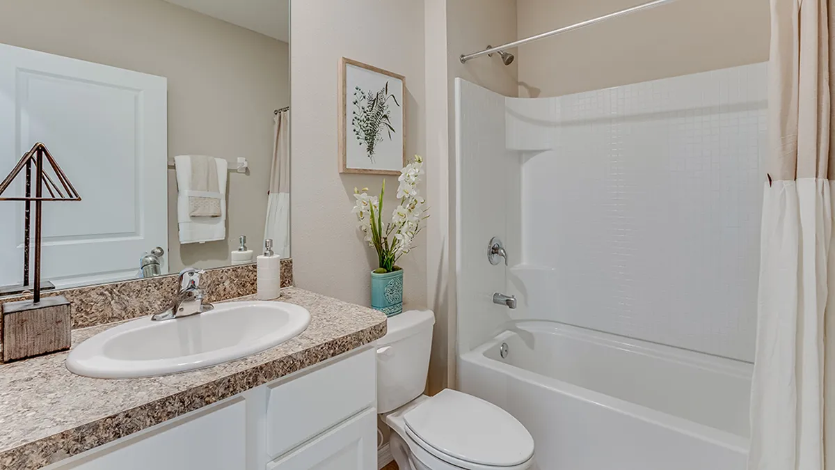 A clean bathroom with a white sink, marble countertop, toilet, and white bathtub-shower combo. A plant and framed artwork decorate the space. Towels hang on the wall, and a beige shower curtain is pulled to the side.