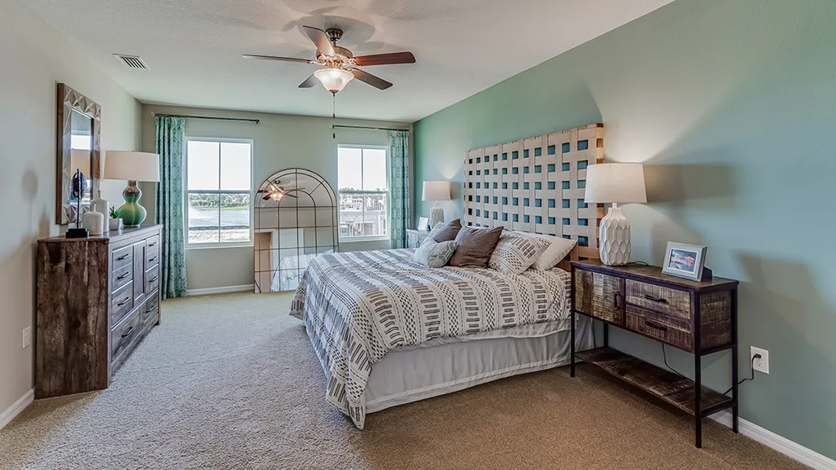 A spacious bedroom with a large bed, patterned bedding, two bedside tables with lamps, a wooden dresser, a fan on the ceiling, and two large windows letting in natural light.