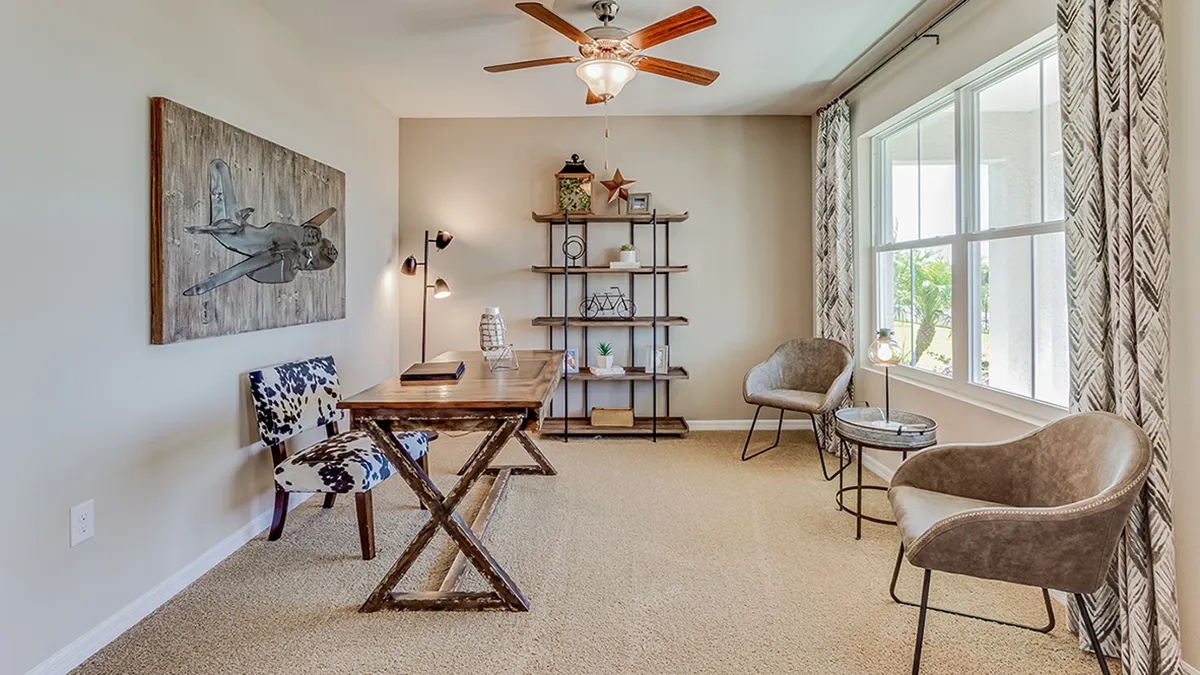 A home office with a wooden desk, patterned chair, modern taupe chairs, a metal shelf with decor, wall art of an airplane, ceiling fan, and large windows with patterned curtains letting in natural light.