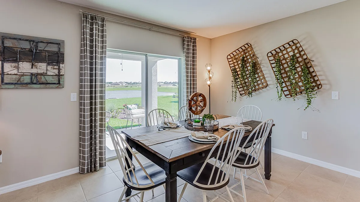 A dining room with a dark wooden table, six mismatched chairs, striped curtains, and wall-mounted planters containing greenery. Large windows reveal a view of a grassy yard and water outside.