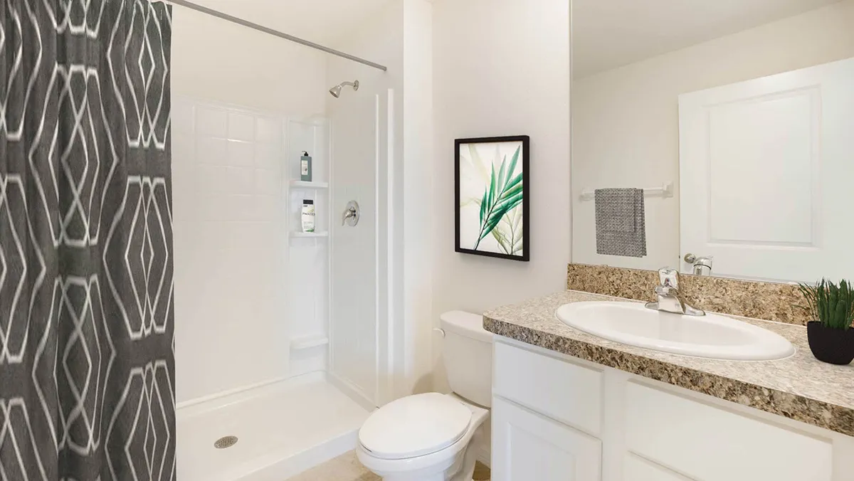 Modern bathroom with white walls, a walk-in shower with geometric-patterned dark gray curtain, white toilet, granite countertop with sink, a towel on the rack, and framed green leaf art on the wall.