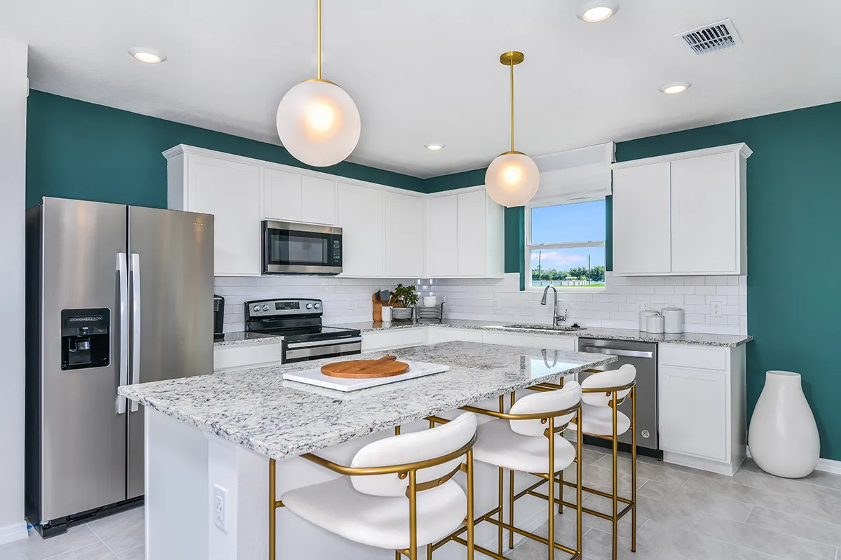 Modern kitchen with white cabinets, stainless steel appliances, marble countertop island with four white and gold bar stools, teal accent walls, pendant lights, and a large window.