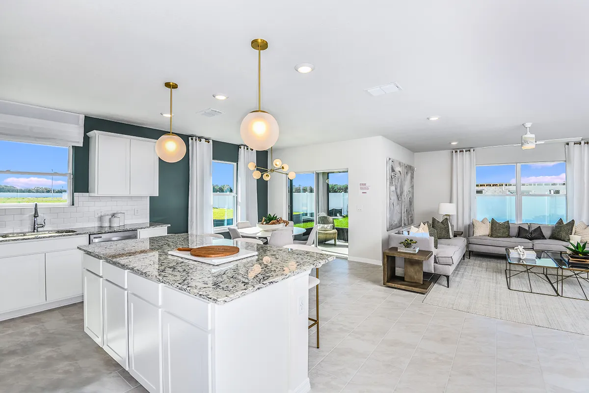 Modern open-concept kitchen and living room with white cabinets, granite countertops, pendant lights, a cozy gray sectional sofa, large windows letting in natural light, and light-colored tiled flooring.