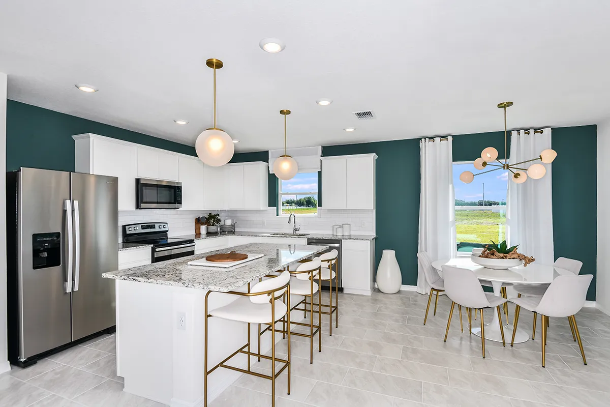 Modern kitchen with white cabinets, stainless steel appliances, granite countertops, and an island with gold-accented stools. Adjacent dining area features a round table, white chairs, and large windows with white curtains.