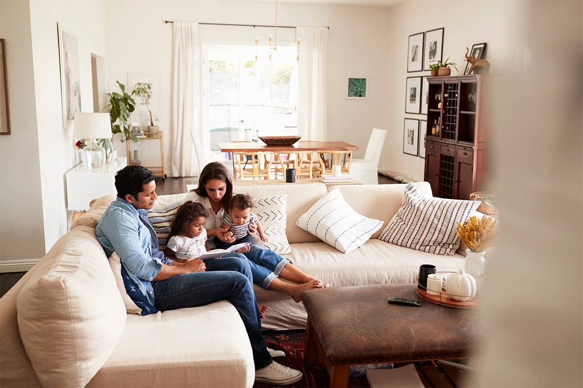 A family of four sits together on a beige sofa in a bright living room. The parents read a book to their two young children. The room is decorated with framed art, cushions, and a dining table in the background.