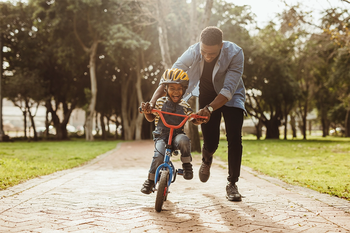 An adult helps a smiling child learn to ride a bike on a park path. The child wears a yellow helmet and grips the handlebars, while the adult runs alongside, holding the bike steady. Trees and grass line the pathway.
