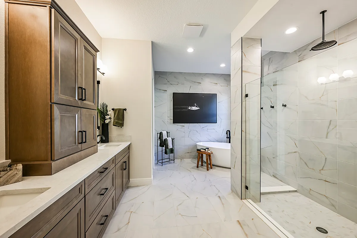 Modern bathroom with wood cabinets, double sinks, a glass-enclosed shower, and a freestanding bathtub against a marble-tiled wall. Decor includes towels, a plant, and a black-and-white wall art piece.