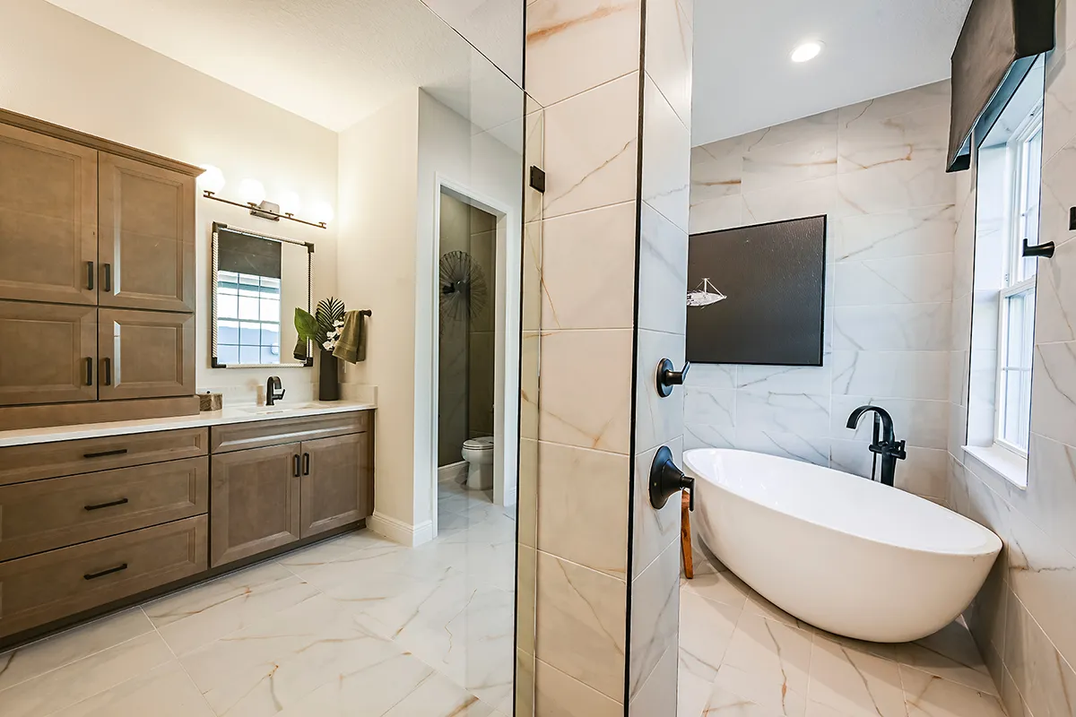 Modern bathroom with light wood cabinets, a large mirror, and vanity on the left; a freestanding white bathtub with black fixtures near a window on the right; and a glass-enclosed shower in the center.