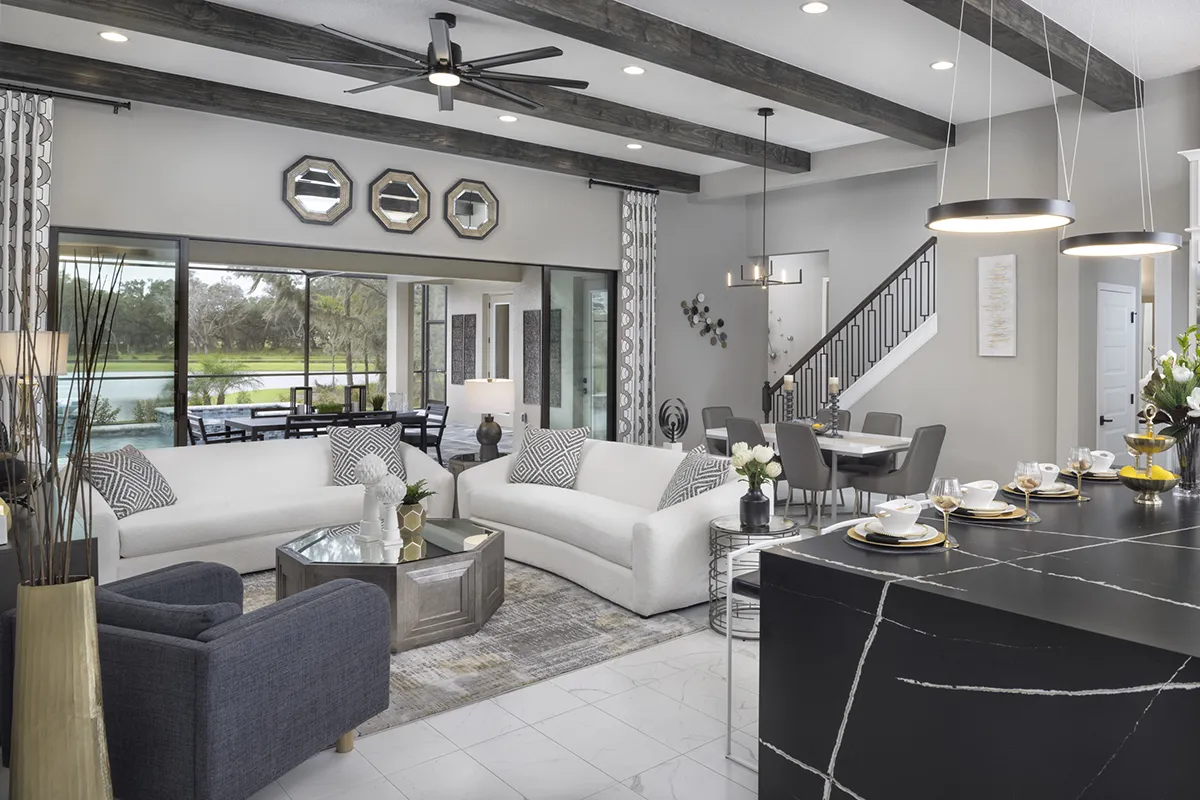 Modern open-concept living room and kitchen with white sofas, dark armchair, glass coffee table, and sleek black marble island; large windows overlook a patio and greenery outside.