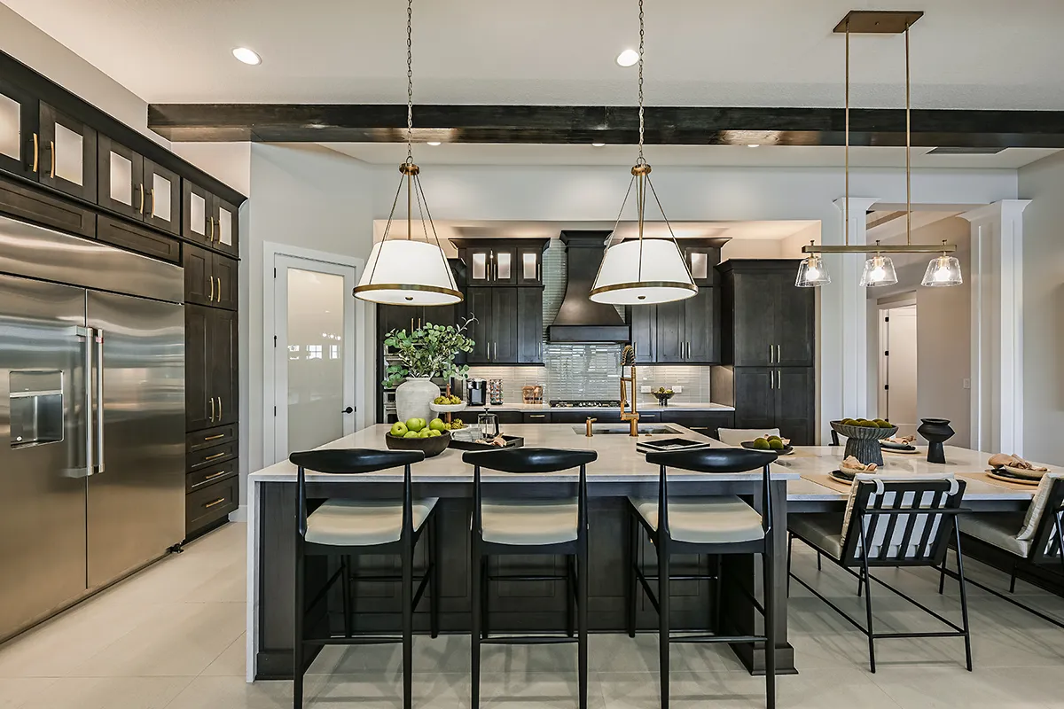 Modern kitchen with dark cabinets, stainless steel appliances, a large island with four chairs, pendant lighting, and decorative items like a fruit bowl and vase on the counter.
