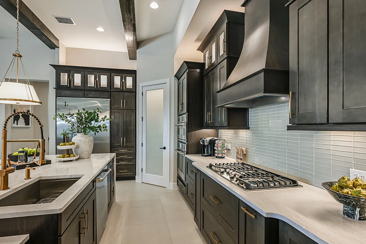 Modern kitchen with dark wood cabinets, white countertops, stainless steel appliances, a gas cooktop, a farmhouse sink, glass tile backsplash, and decorative accents like fruit bowls and plants.