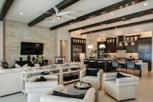 Modern open-concept living room and kitchen with beige armchairs, a sectional sofa, dark wood beams, stone accent wall with TV, and a kitchen island with barstools under pendant lights.