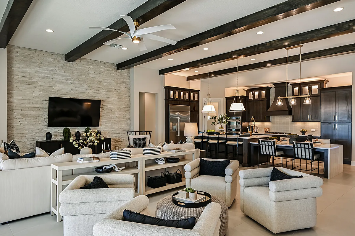 Modern open-concept living room and kitchen with beige armchairs, a sectional sofa, dark wood beams, stone accent wall with TV, and a kitchen island with barstools under pendant lights.
