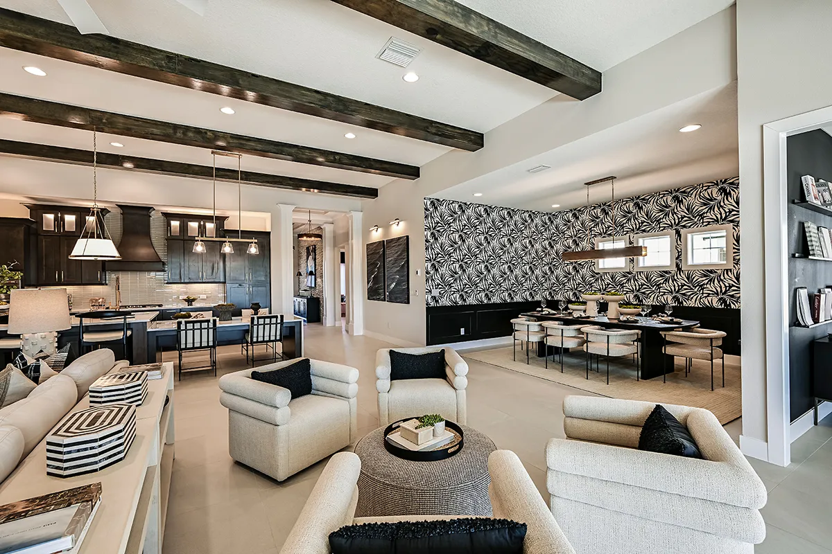 Modern open-concept living space with exposed ceiling beams, neutral-toned sofas and chairs, black-and-white patterned wallpaper in the dining area, and a sleek kitchen with bar seating in the background.